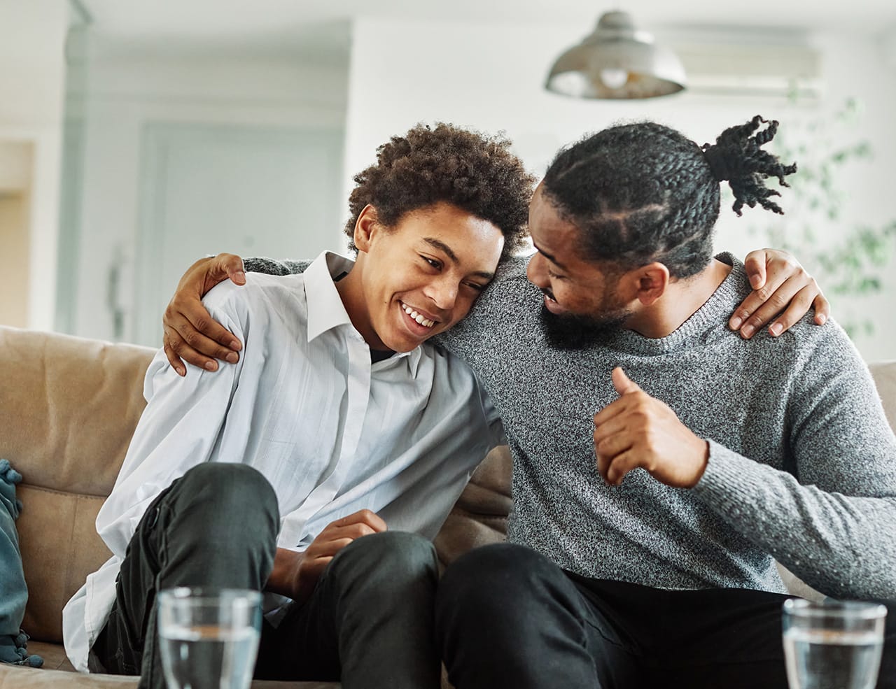 A father and teenage son sit on a couch indoors, smiling and sharing a warm moment. The father is wearing a gray sweater has their arm around his son, who is wearing a white shirt. They are leaning in close, appearing supportive and happy. Glasses of water sit on the table in front of them, and the background shows a cozy, well-lit room.