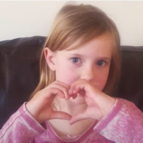 A young girl with light brown hair is sitting on a couch, wearing a pink long-sleeve shirt. She is holding her hands together in the shape of a heart and looking directly at the camera with a gentle expression.