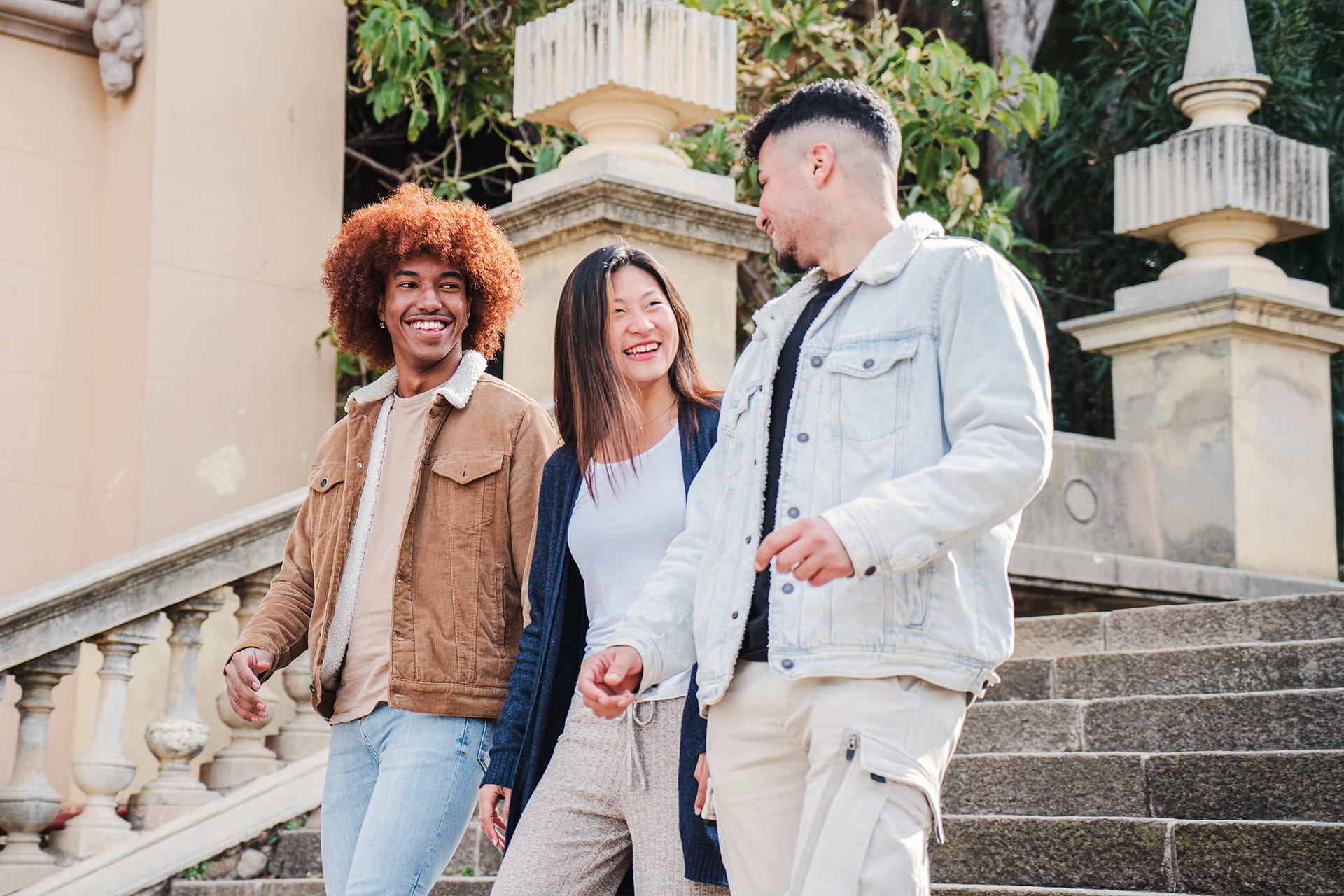 Three friends are walking together outdoors on a stone staircase, smiling and chatting. The person on the left is wearing a brown jacket and jeans, the person in the middle is wearing a navy cardigan and light pants, and the person on the right is wearing a light denim jacket and light cargo pants. There are decorative stone railings and greenery in the background.
